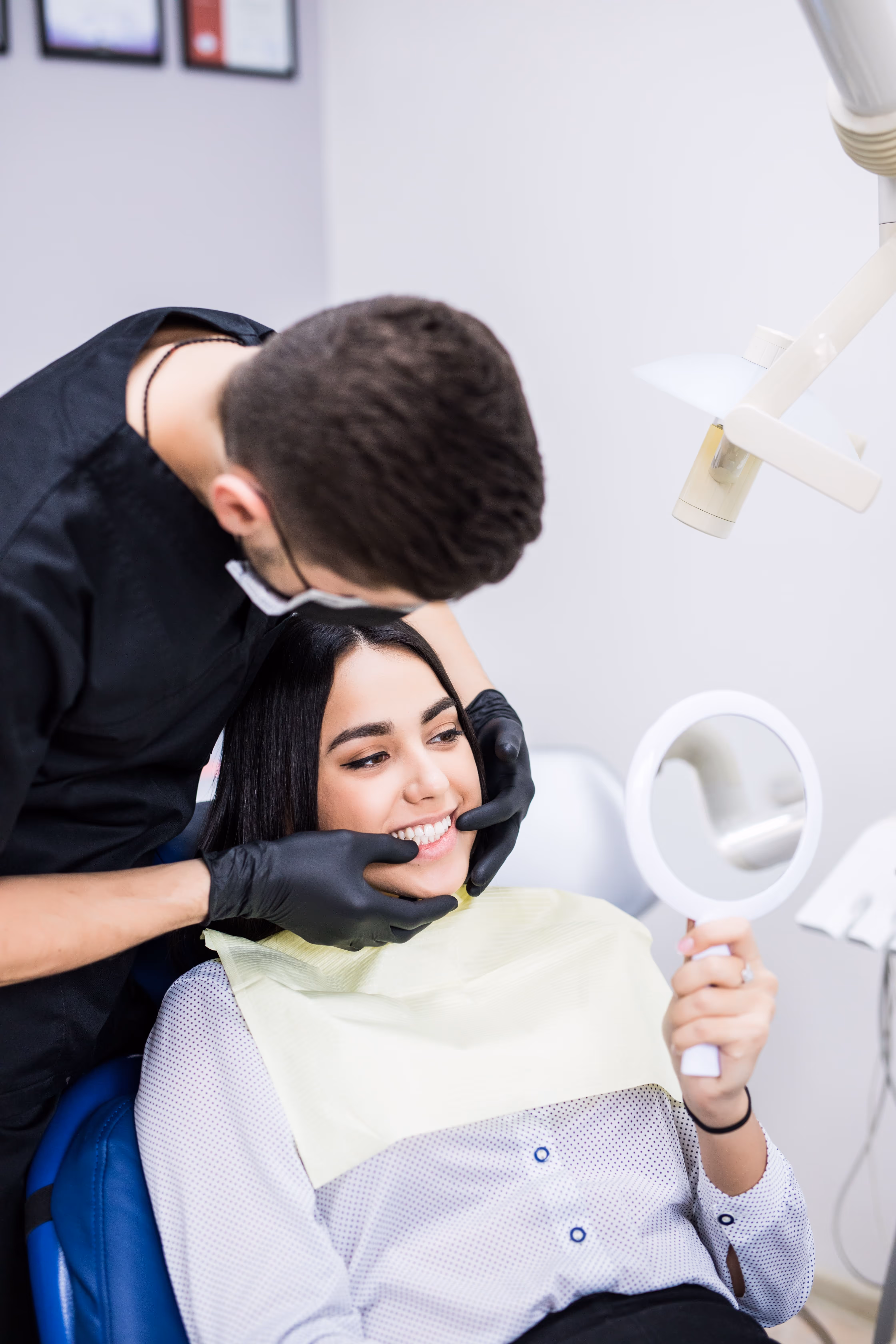 Dentist wearing black gloves examining smiling patient's teeth while she holds a hand mirror.