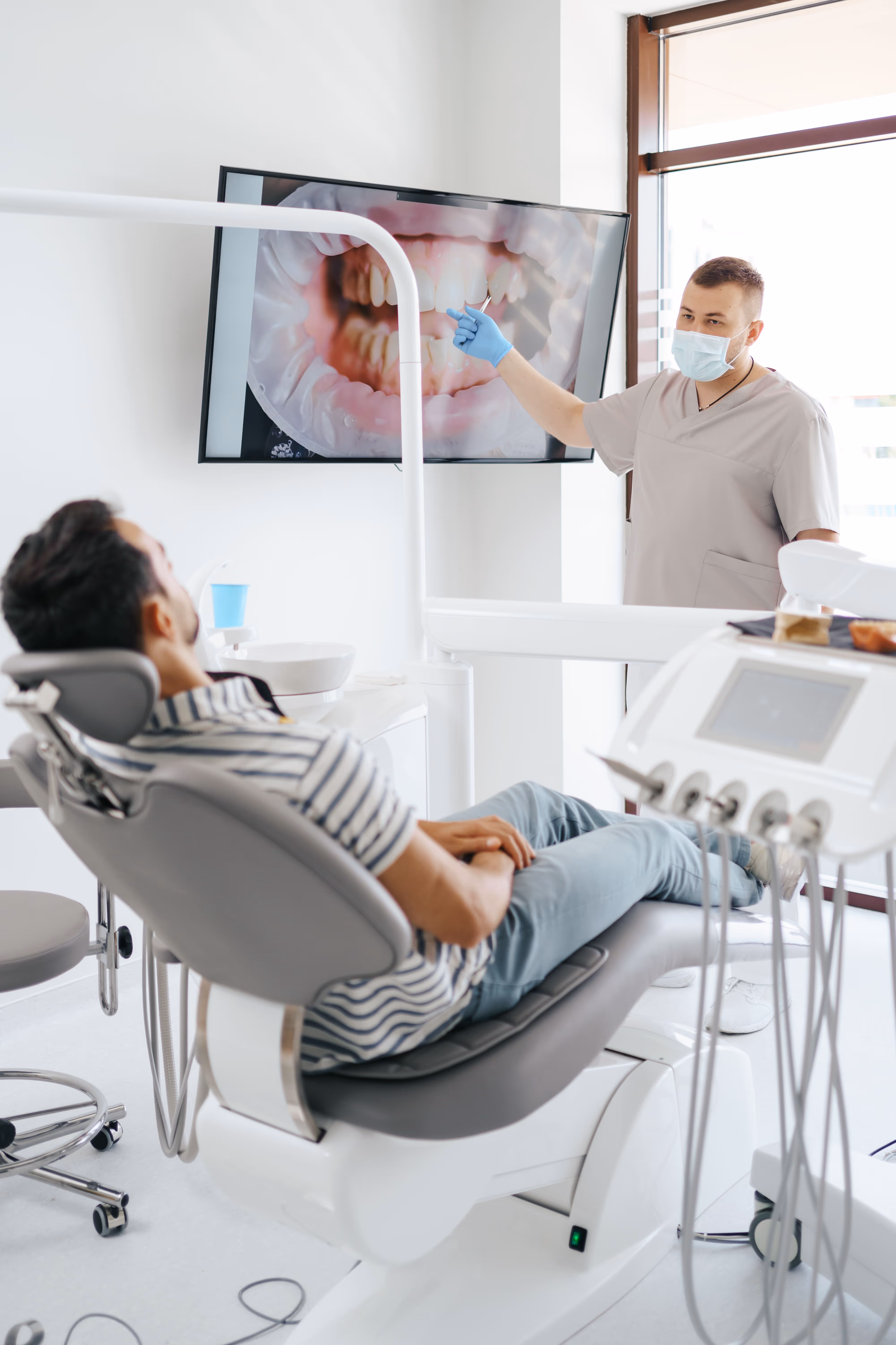 Dentist wearing mask and gloves explains dental condition to a male patient sitting in a dental chair using a large screen showing close-up of teeth.