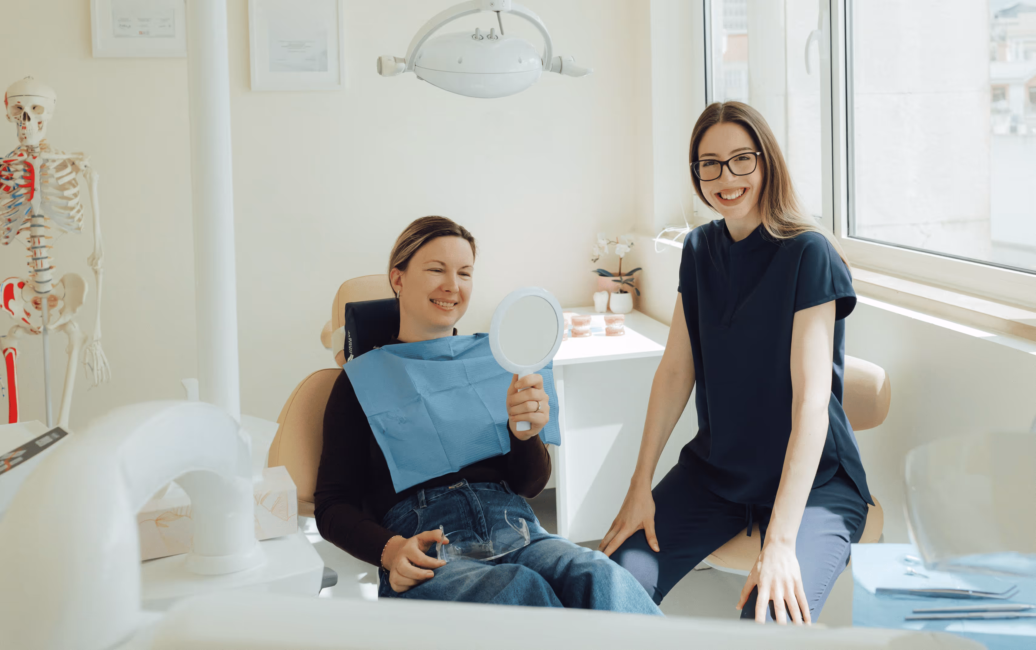 Smiling female patient holding a hand mirror in a dental clinic with a female dentist sitting beside her.