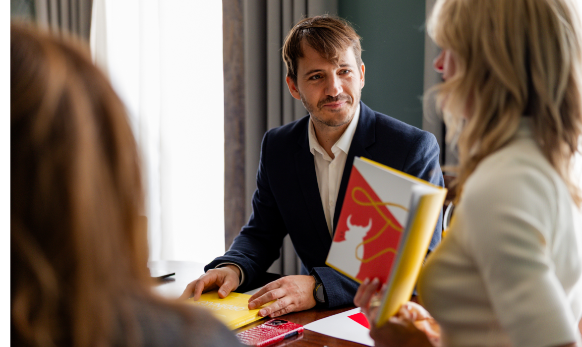 A man in a dark blazer sitting at a table looks at a woman holding open a colorful book with red and yellow designs; another person is partially visible in the foreground.