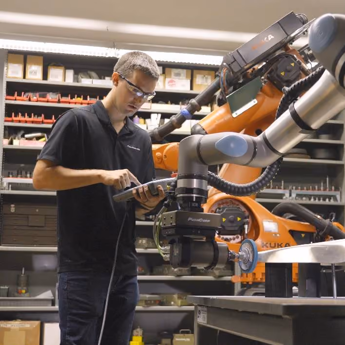 A PushCorp engineer with a teach pendant programming an orange KUKA robot fitted with a PushCorp force compliance device and spindle