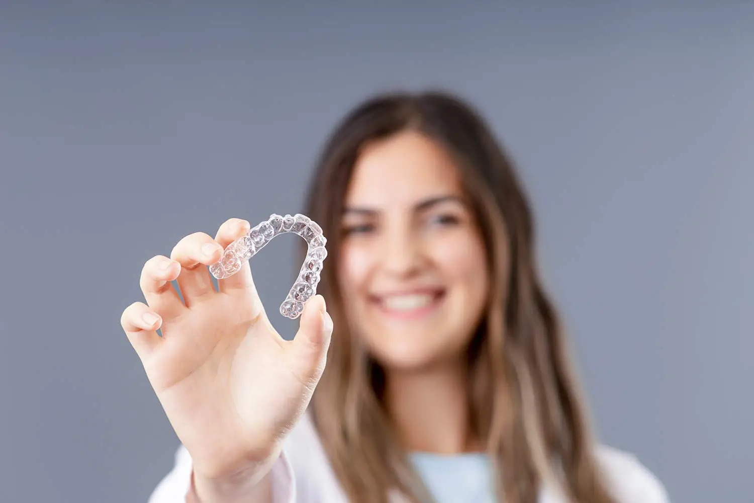 Smiling woman holding a clear dental aligner in front of a gray background.
