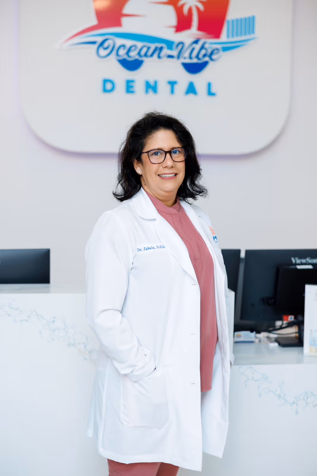 Female dentist wearing glasses and a white coat standing in front of an Ocean-Vibe Dental sign and reception desk.
