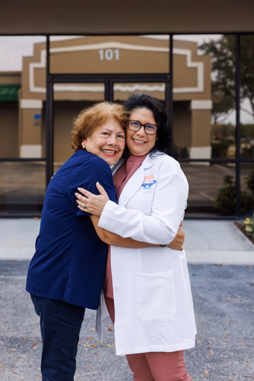 Two women smiling and hugging outside a building with the number 101, one wearing a white dental coat and the other in a blue shirt.