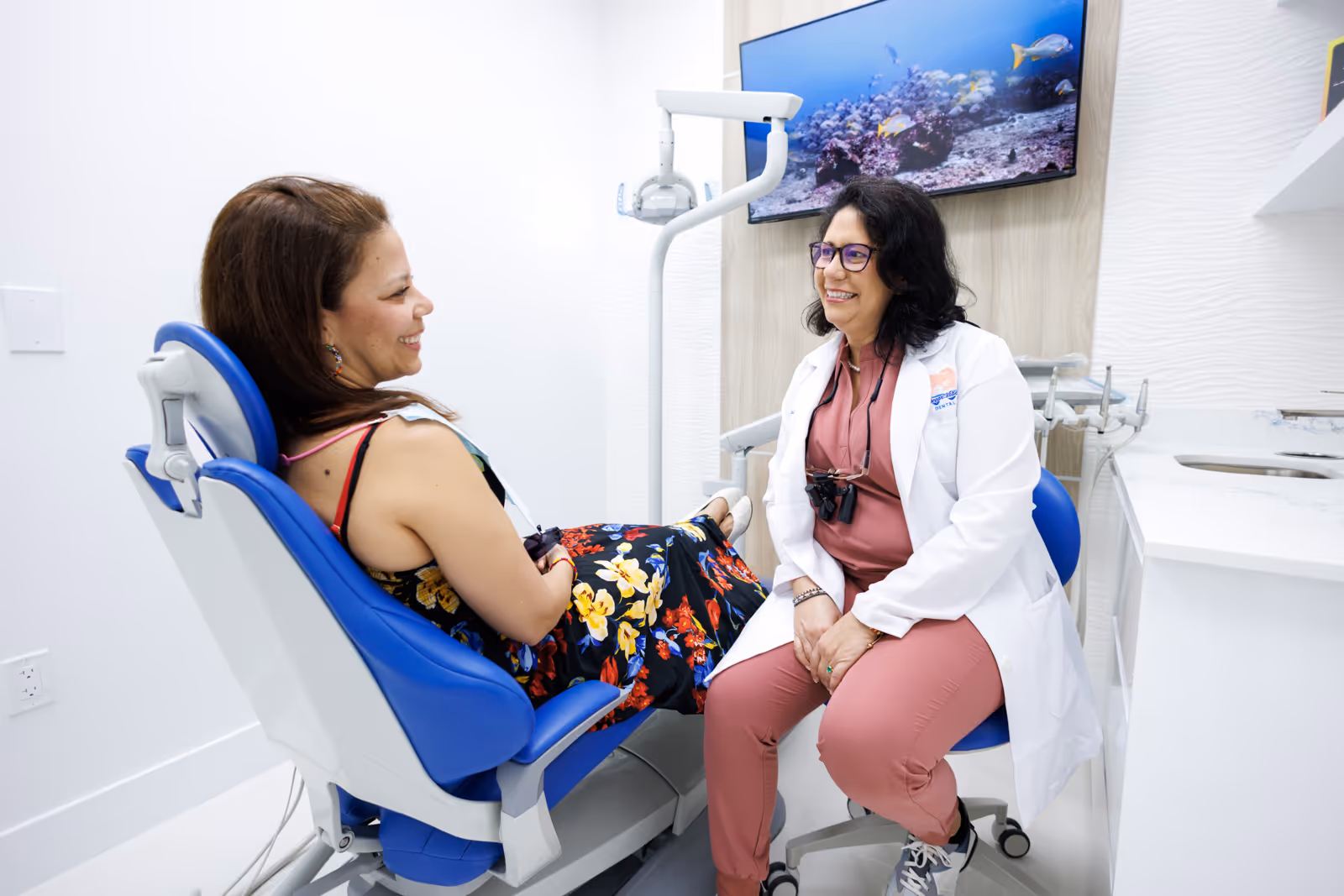 Dentist in white coat smiling and talking with a female patient seated in a dental chair.