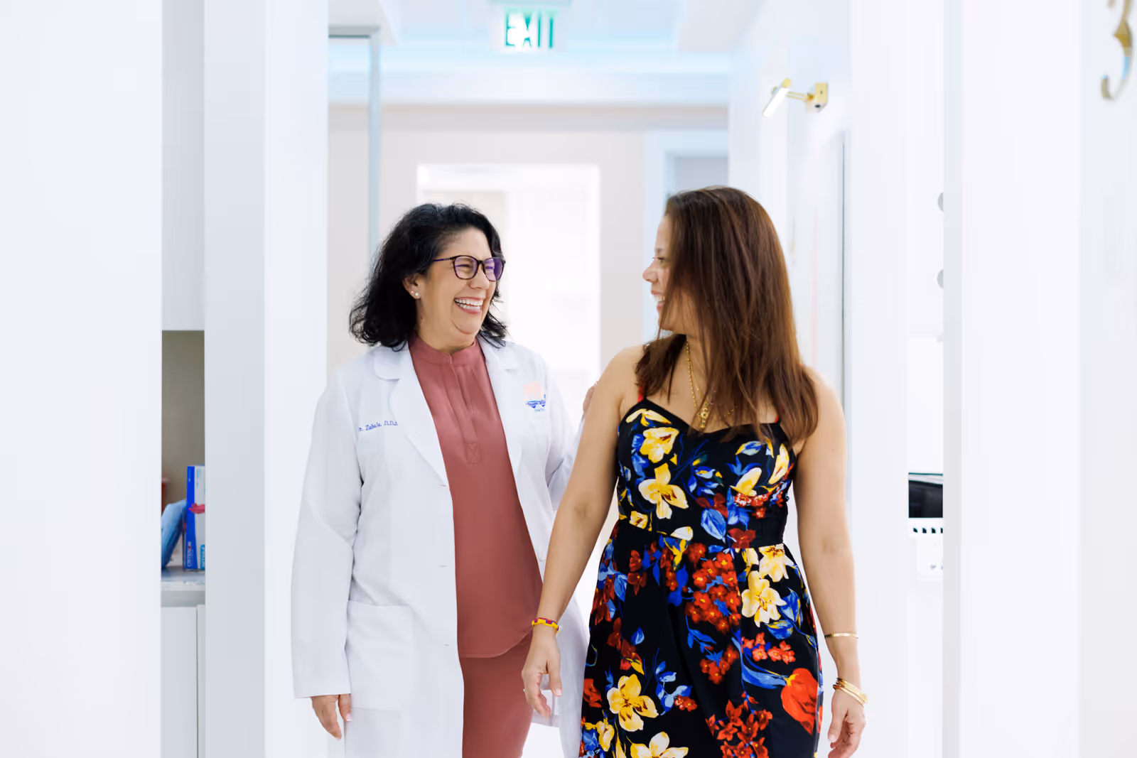 Female doctor in white coat smiling and walking with a woman in a black floral dress in a bright hallway.