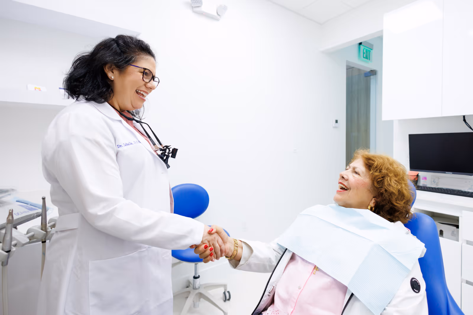 Female dentist in white coat shaking hands and smiling with an elderly female patient sitting in a dental chair.