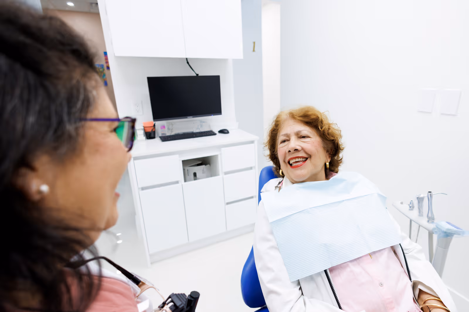 Smiling elderly woman sitting in a dental chair wearing a bib, talking to a dentist in a bright dental office.