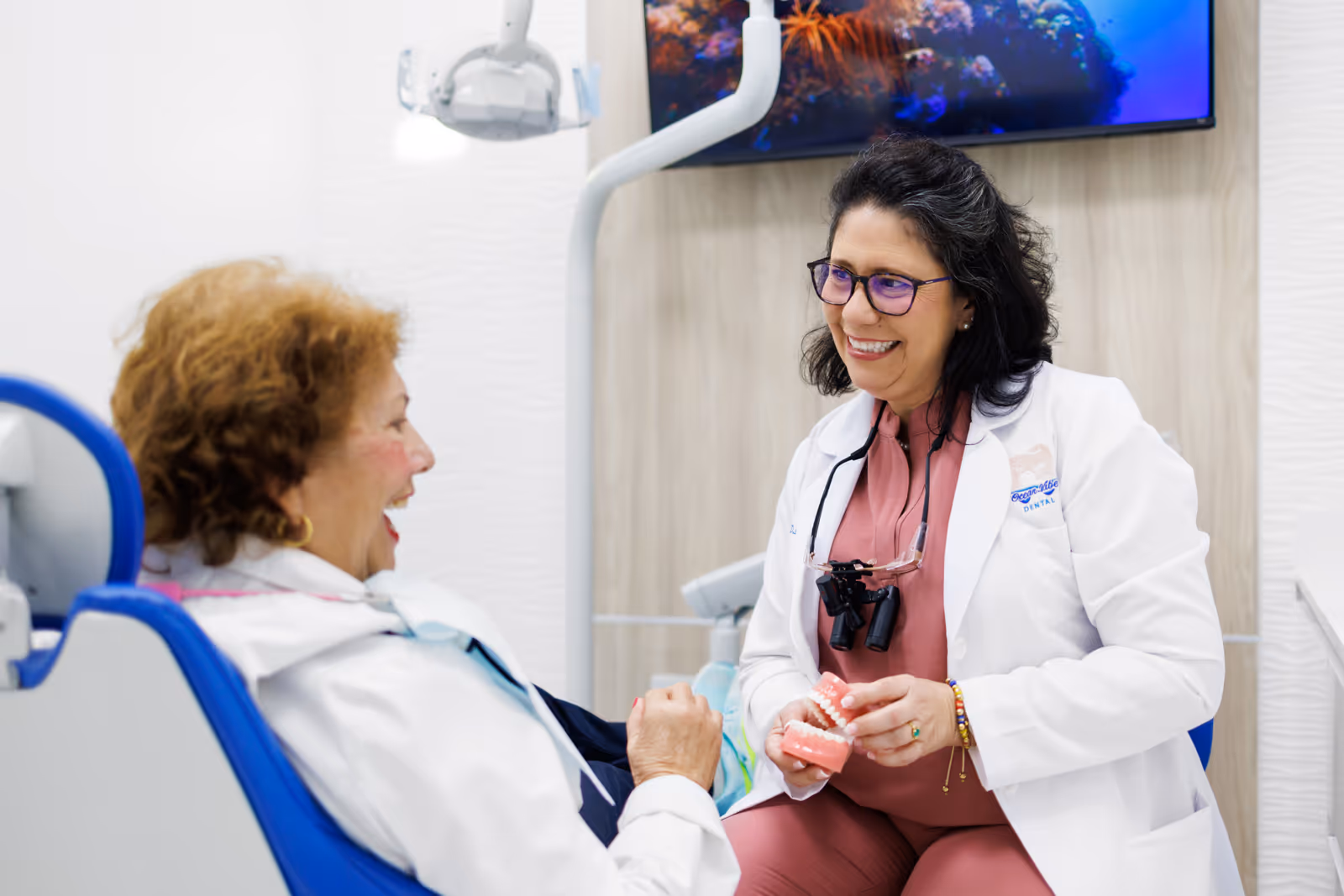 Female dentist in a white coat smiling and holding a dental model while talking to a female patient sitting in a dental chair.