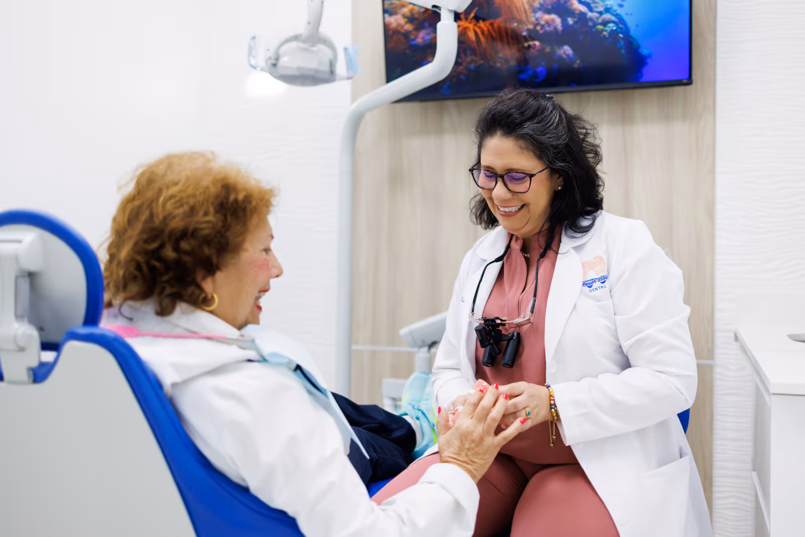 Dentist smiling and holding hands with a female patient seated in a dental chair in a modern clinic.