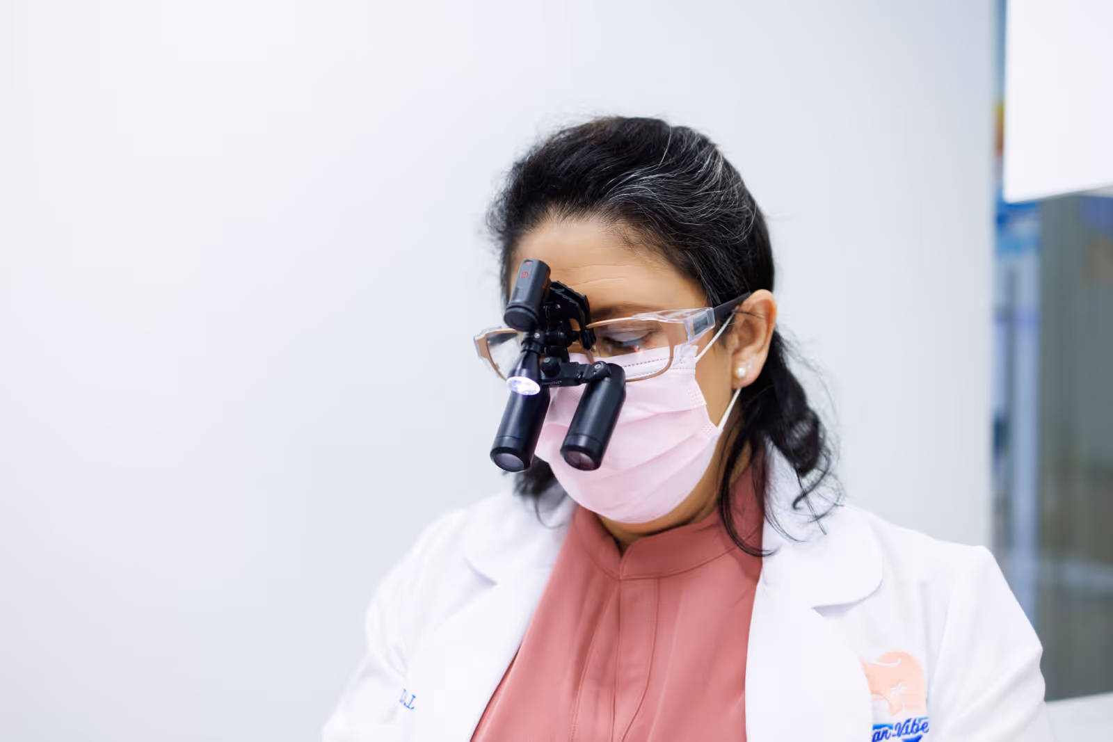 Female dentist wearing magnifying loupes, protective glasses, a pink mask, and a white coat in a clinical setting.