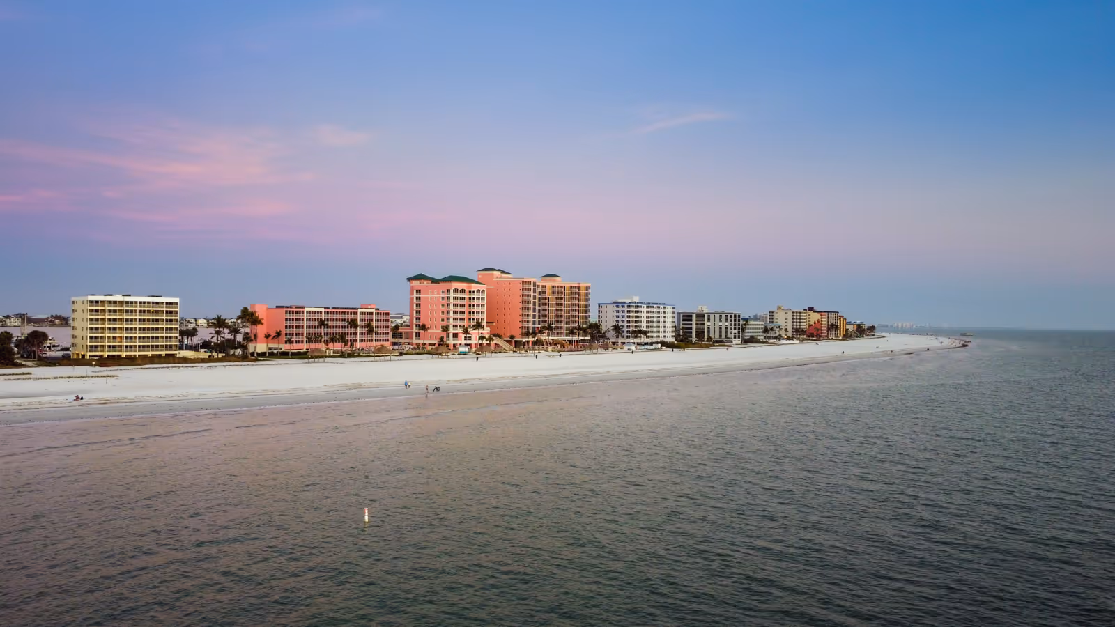 Panoramic view of a coastal beachfront with white sand, lined by colorful resort buildings under a pastel sunset sky.