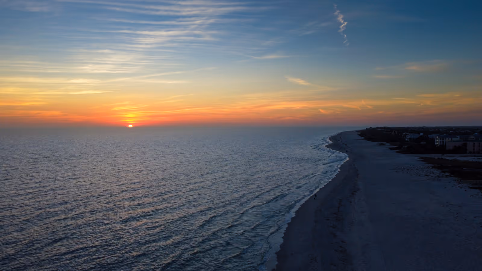 Sunset over ocean waves with a long sandy beach and distant buildings along the shore.