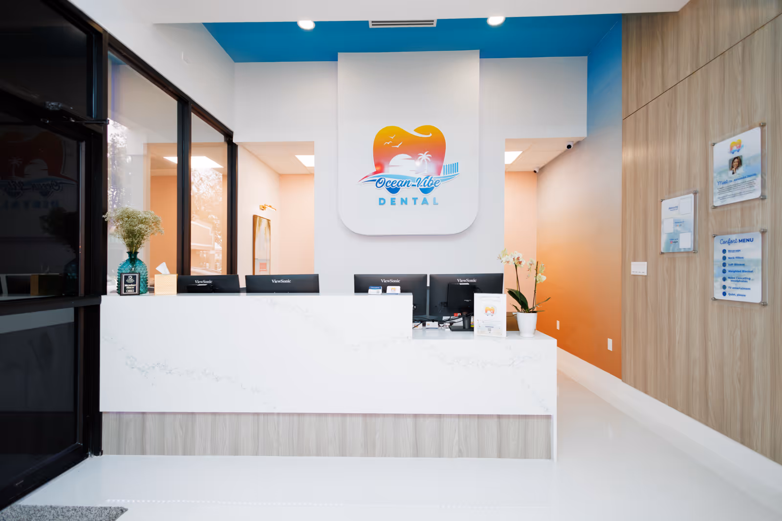 Modern dental reception area with white counter, four computer monitors, floral decorations, and Ocean Vibe Dental logo on the wall.