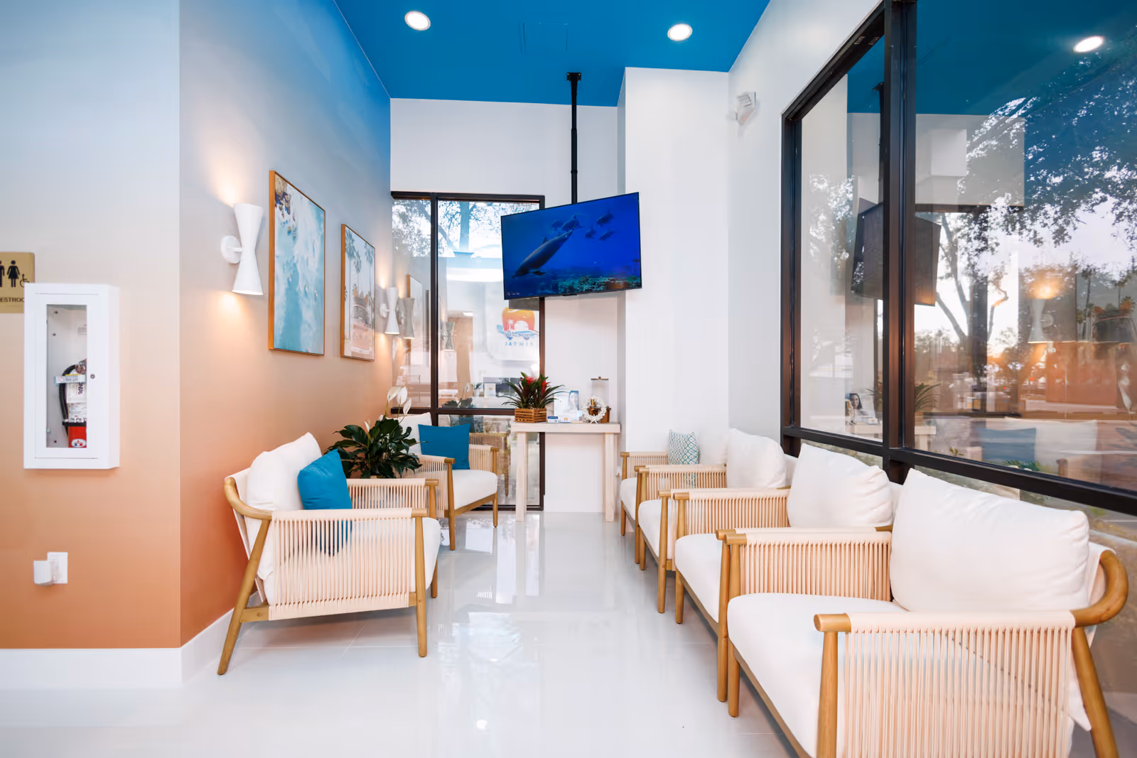 Modern waiting room with wooden framed white cushioned chairs, blue accent pillows, large windows, and a TV mounted on the wall.
