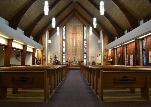 Interior shot of the sanctuary at St. John's Lutheran Church in Algoma, Wisconsin, with wooden pews facing a large cross and stained glass windows behind the altar. 