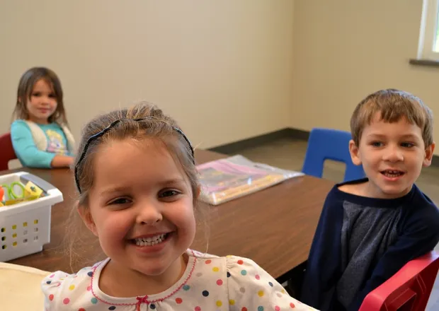 Three smiling children seated at a table in a classroom, with a bin of craft supplies beside them.