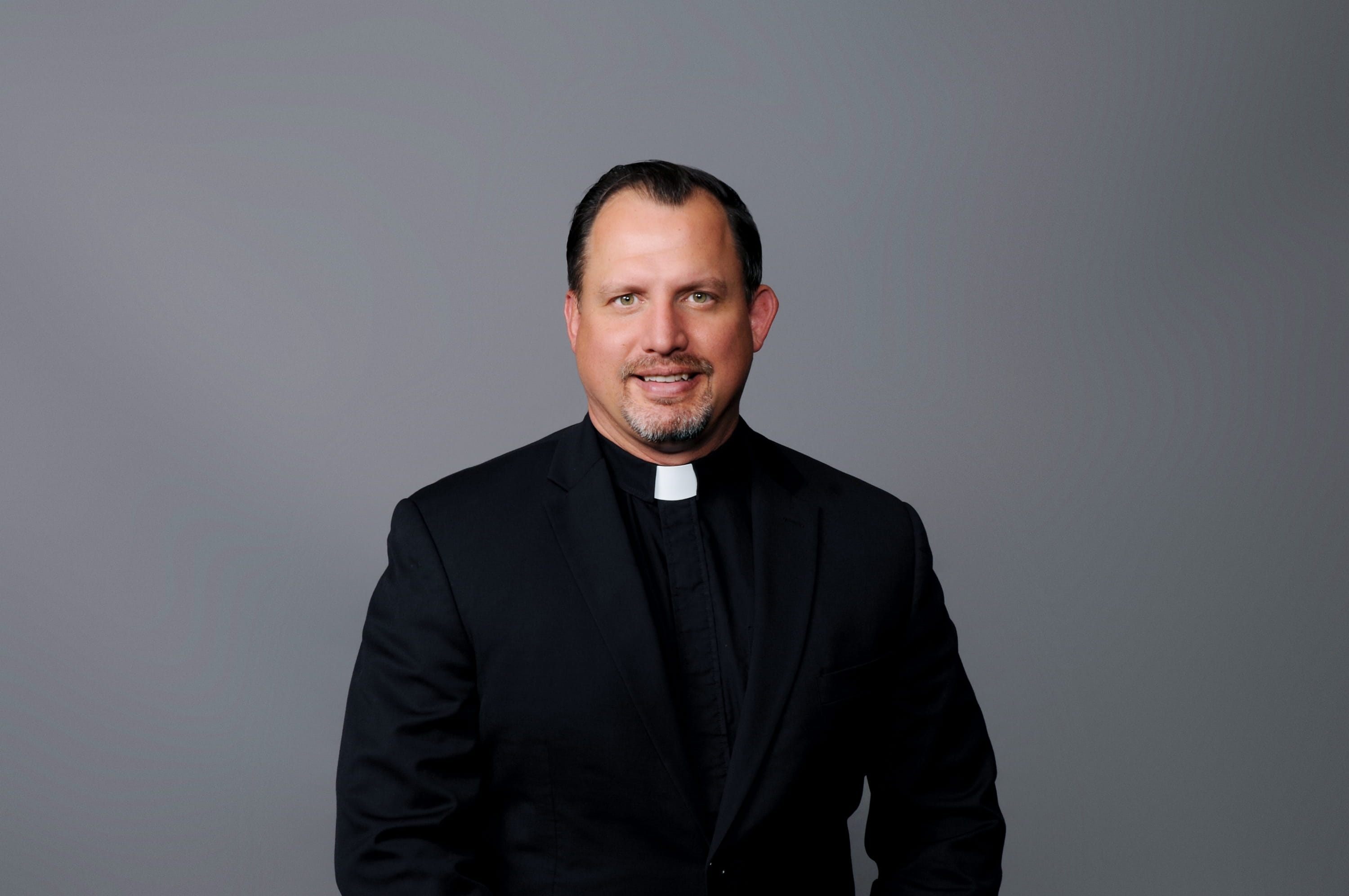 Headshet of Pastor Chris Jackson of St. John's Lutheran Church in Algoma, Wisconsin. He wears a black clerical shirt and suit jacket in front of a grey background. 