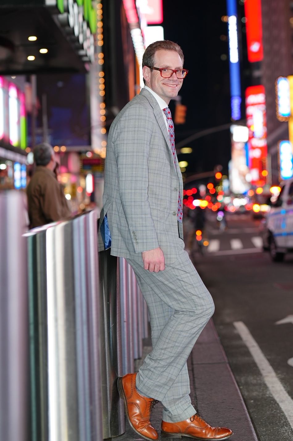 Austin Parker in a gray plaid suit leaning on a sidewalk railing in Times Square at night