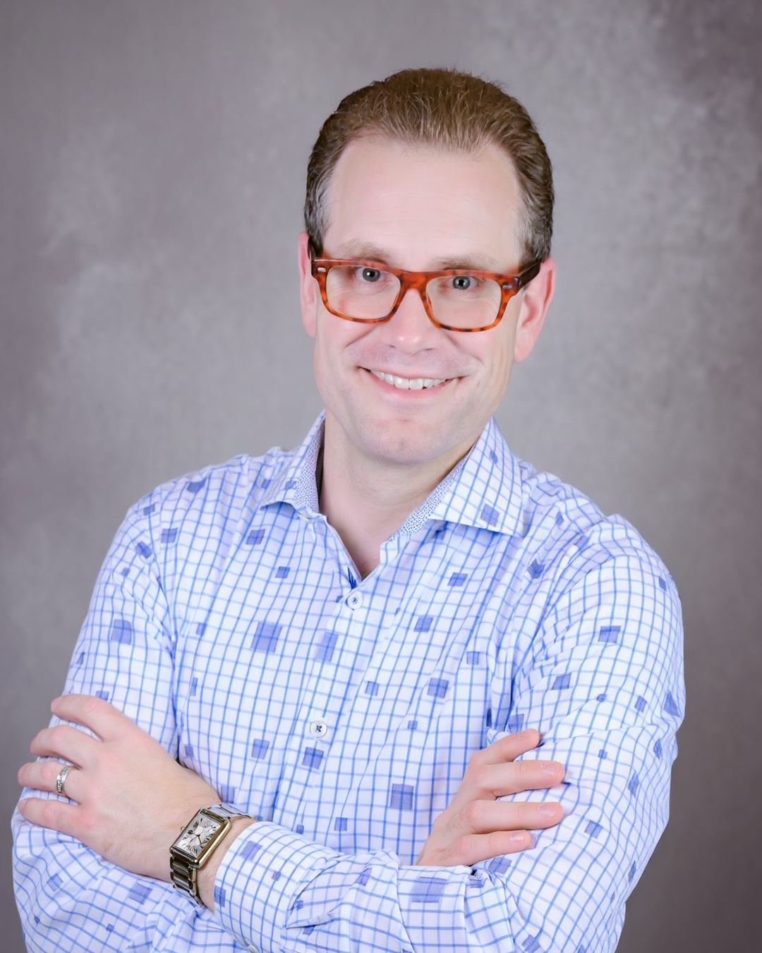 Austin Parker in a blue checked shirt, arms crossed and smiling, against a gray studio backdrop