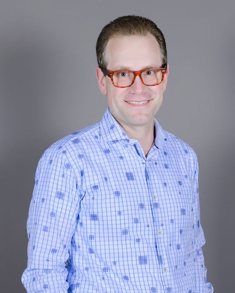 Austin Parker in a blue checked shirt, smiling, against a gray studio backdrop