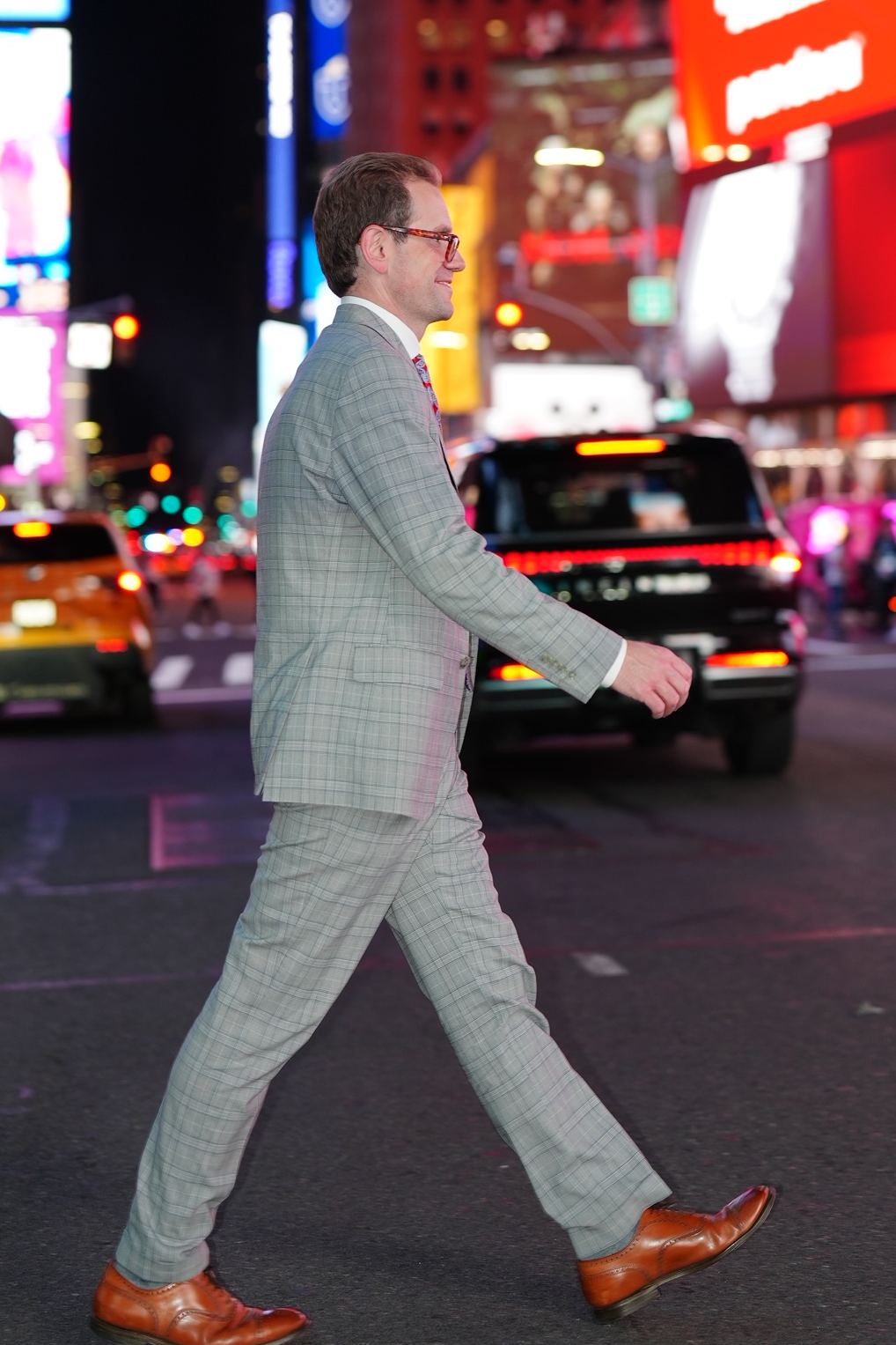 Austin Parker in a gray plaid suit walking across a street in Times Square at night