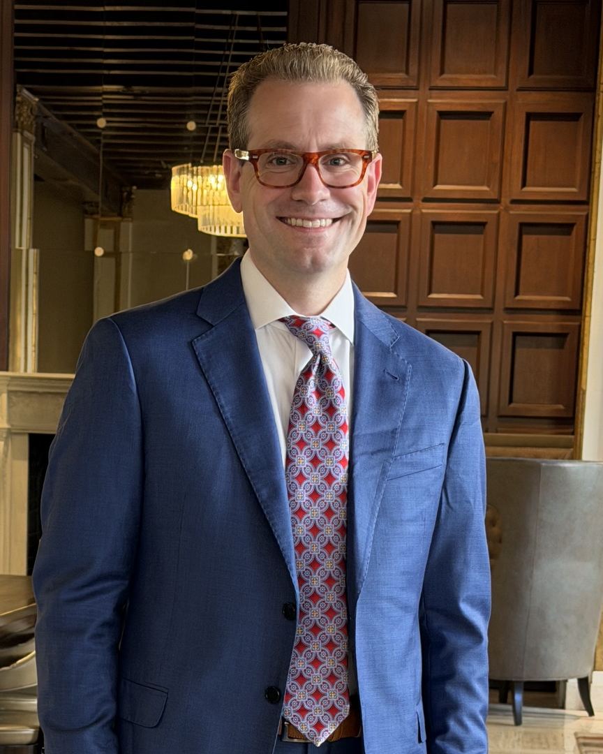 Austin Parker in a blue suit and patterned tie, smiling, in a wood-paneled lounge