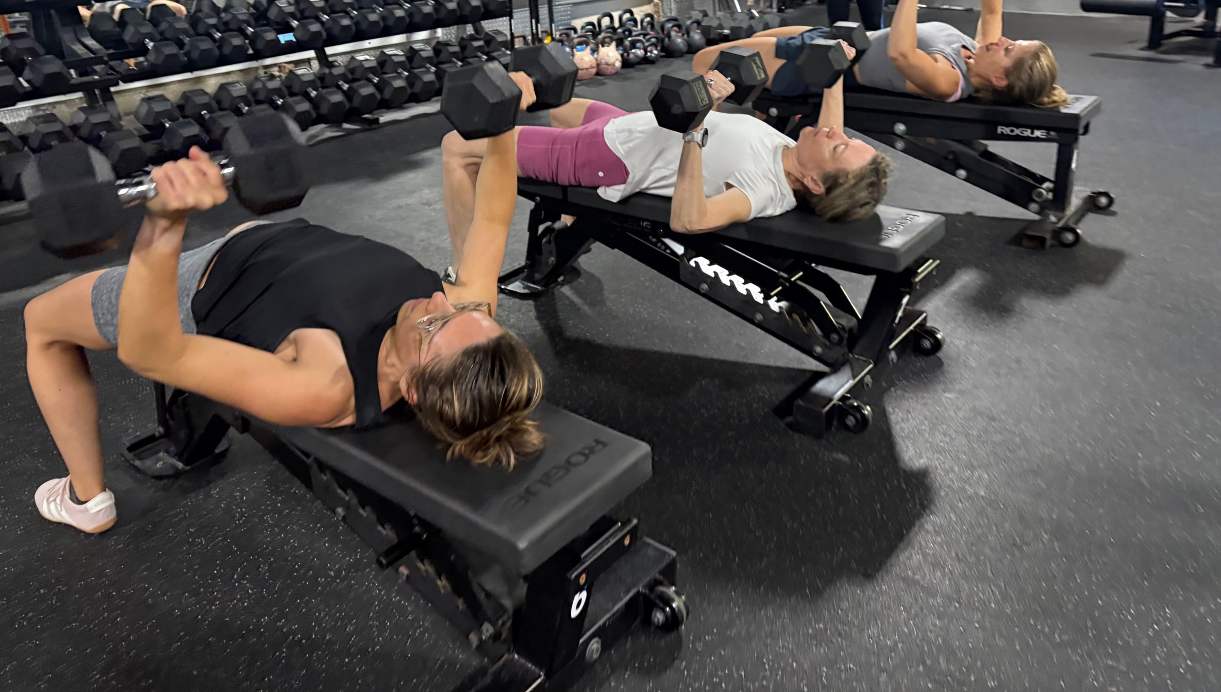 Three women doing dumbbell presses on benches at Three Bears Fitness in Forest Park while a coach looks on