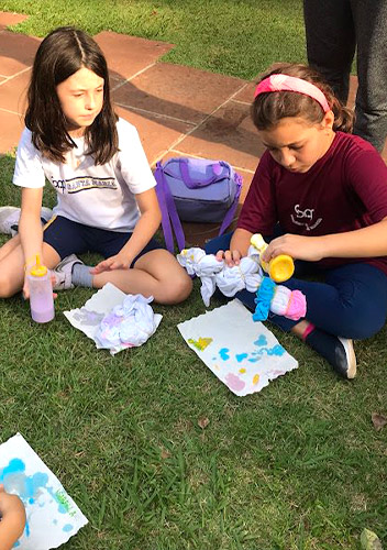 Duas estudantes pintam tecidos e papéis sentadas no gramado da escola