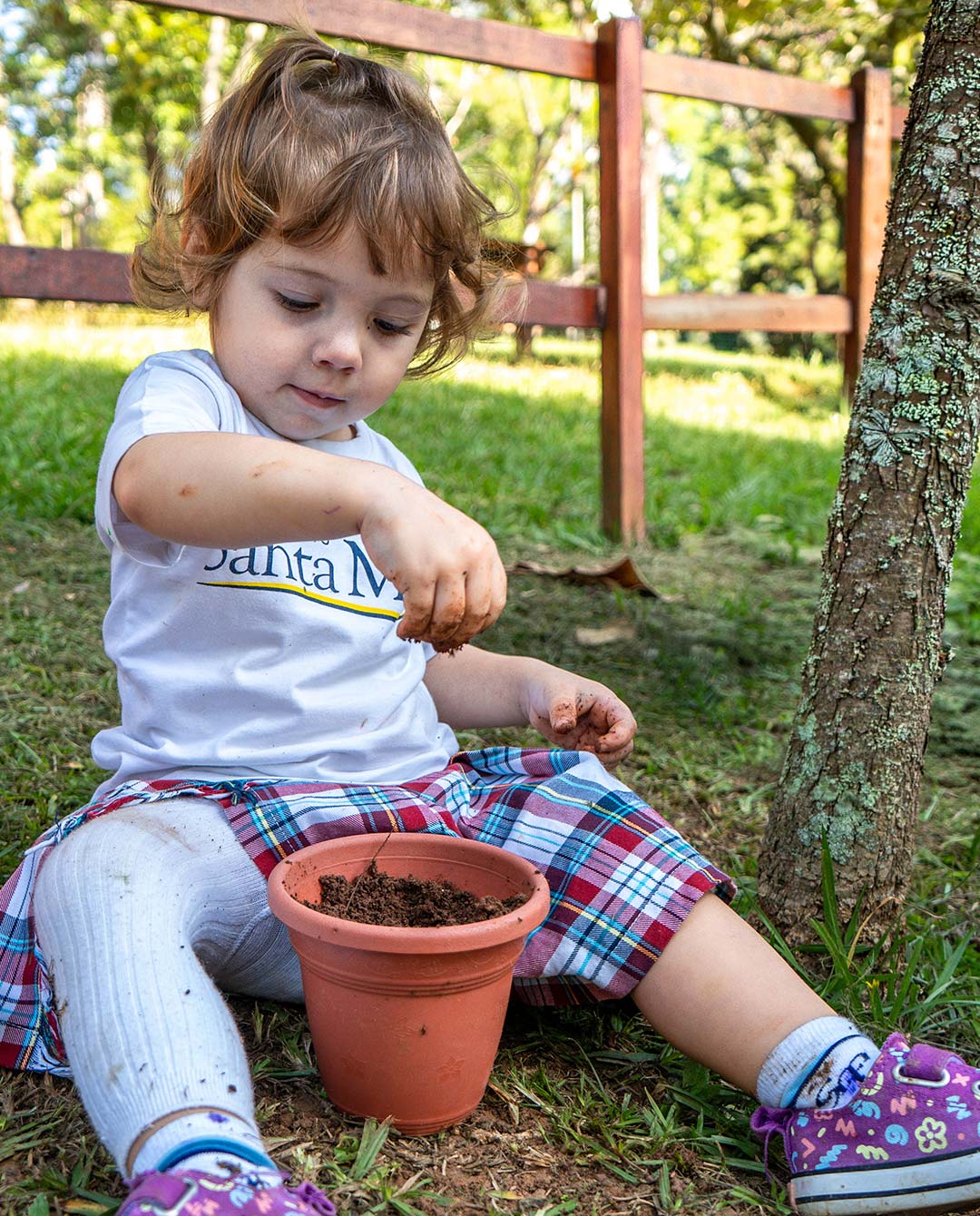 Criança planta em um vaso sentada no gramado ao lado de uma árvore