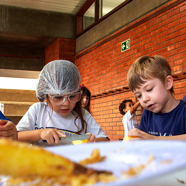 Duas crianças cortam frutas com apoio de utensílios durante atividade culinária