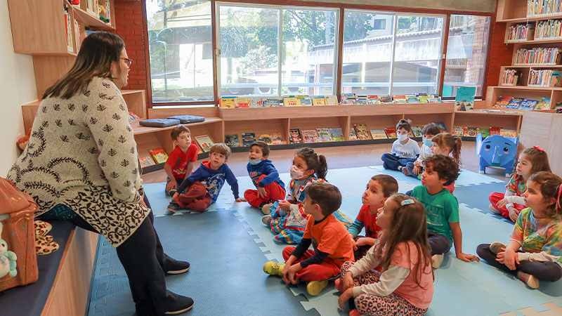 Educadora conta uma história para crianças sentadas no chão da biblioteca infantil