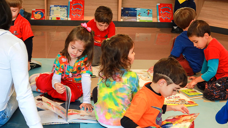 Crianças folheiam livros sentadas no chão da biblioteca infantil