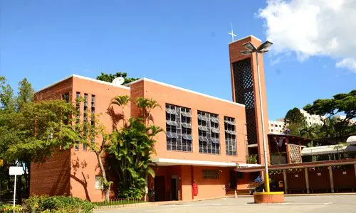 Prédio de tijolos do Ensino Fundamental com torre e cruz sob o céu azul