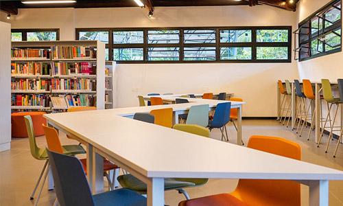 Biblioteca com mesas, cadeiras coloridas, estantes de livros e janelas amplas