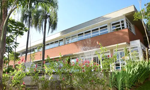 Prédio do Ensino Médio cercado por árvores, palmeiras e jardim florido