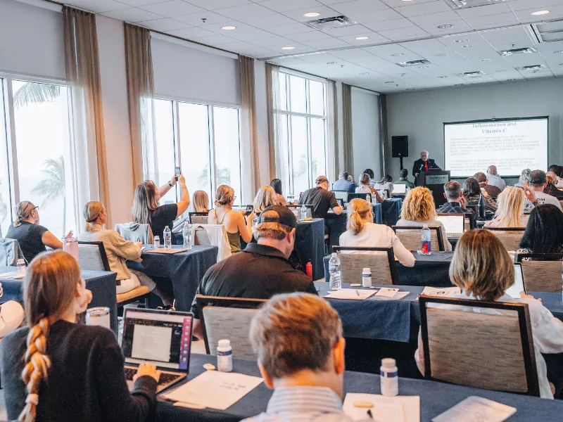 Clinicians at classroom tables take notes while a speaker presents on inflammation and vitamin C