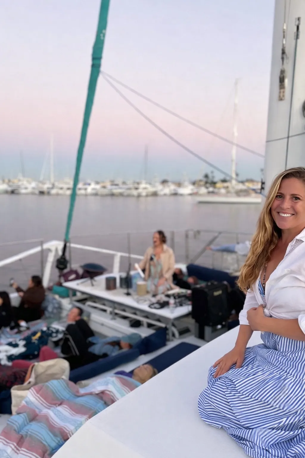 Stephanie smiling on a boat deck at dusk, with a pastel pink sky over the Coronado marina and guests resting on cushions behind her