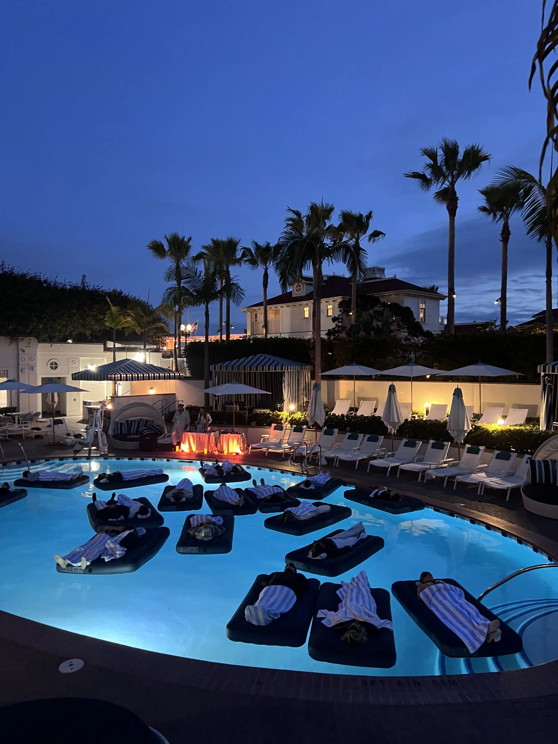 Guests resting on dark floating daybeds in a glowing blue resort pool at night, with palm trees and cabanas in the background