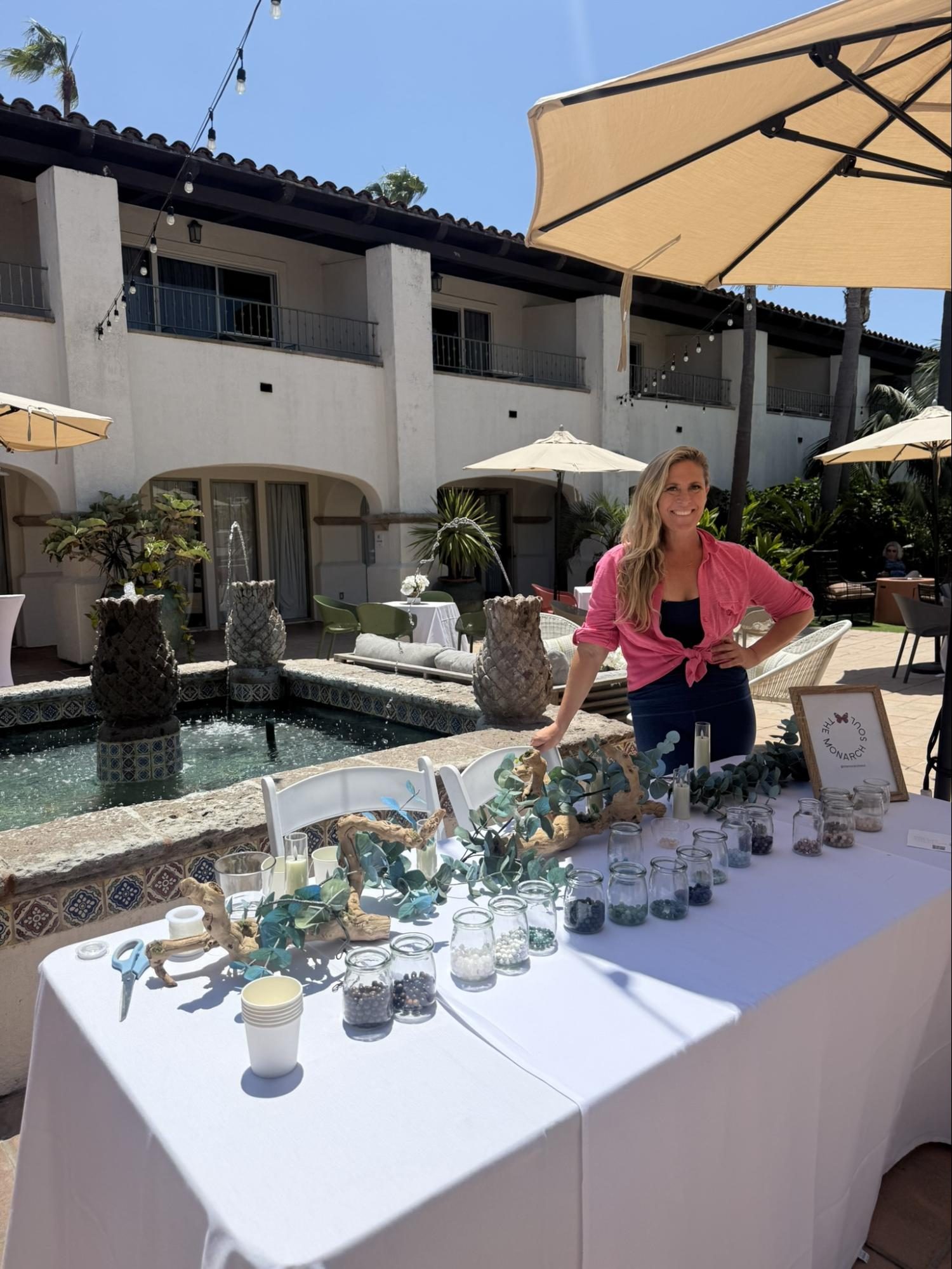 Stephanie smiling at a table of bead jars and eucalyptus in a sunny courtyard with a tiled fountain, under a patio umbrella.