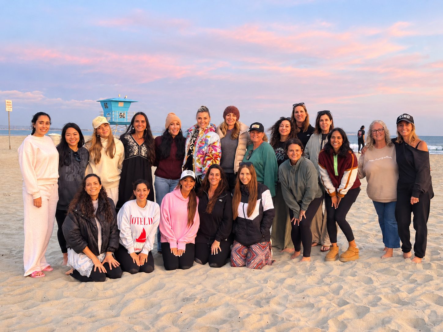 A group of women posing together on the beach at sunset, with a blue lifeguard tower behind them.
