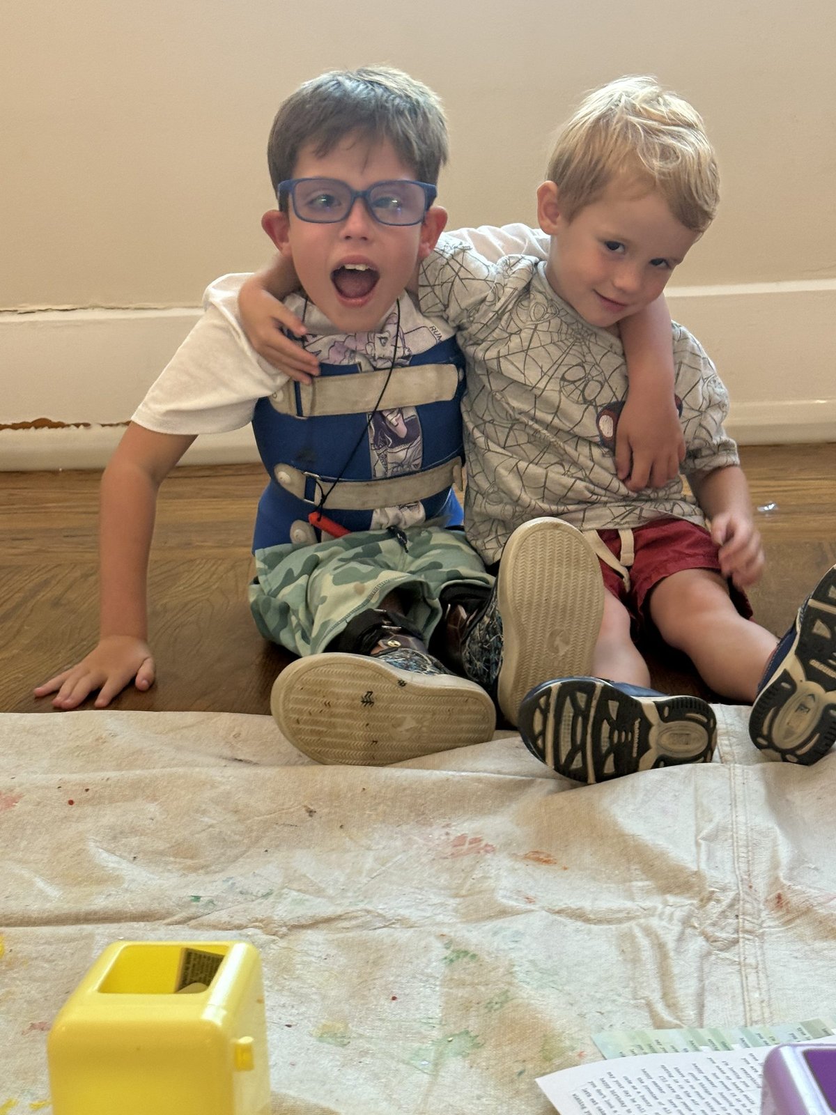 Two young boys hugging and laughing on a paint-splattered drop cloth at Monarch Kids