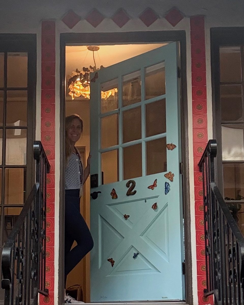 Stephanie smiling in the open doorway of The Monarch Soul studio, a pale blue door decorated with butterflies at the top of red-tiled steps