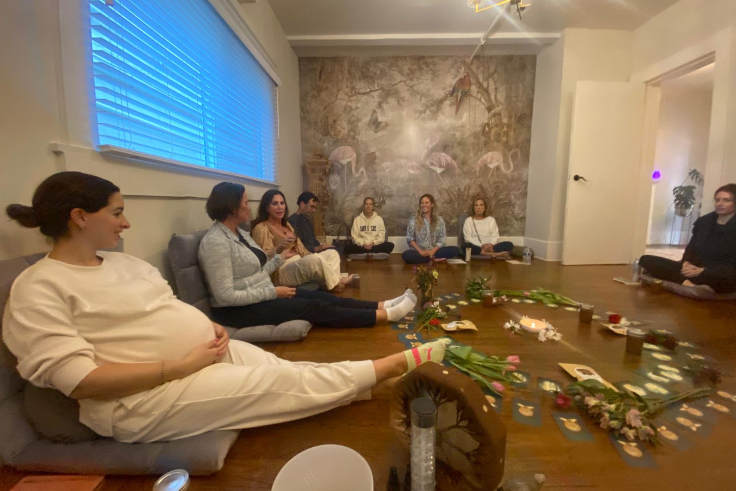A group sitting in a circle on floor cushions in the Monarch Soul studio, around a centerpiece of flowers, candles and cards