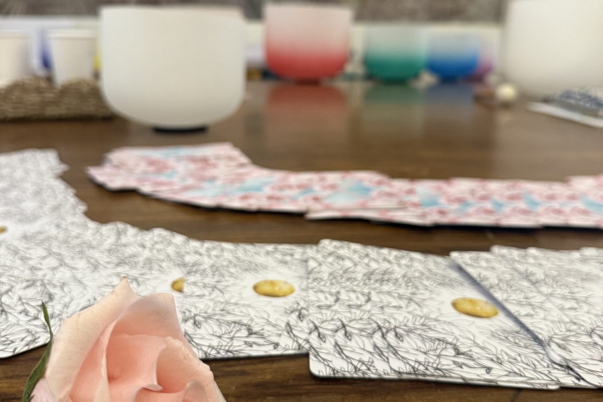 A pale pink rose beside a spread of oracle cards and crystal singing bowls on a wooden table in the Monarch Soul studio