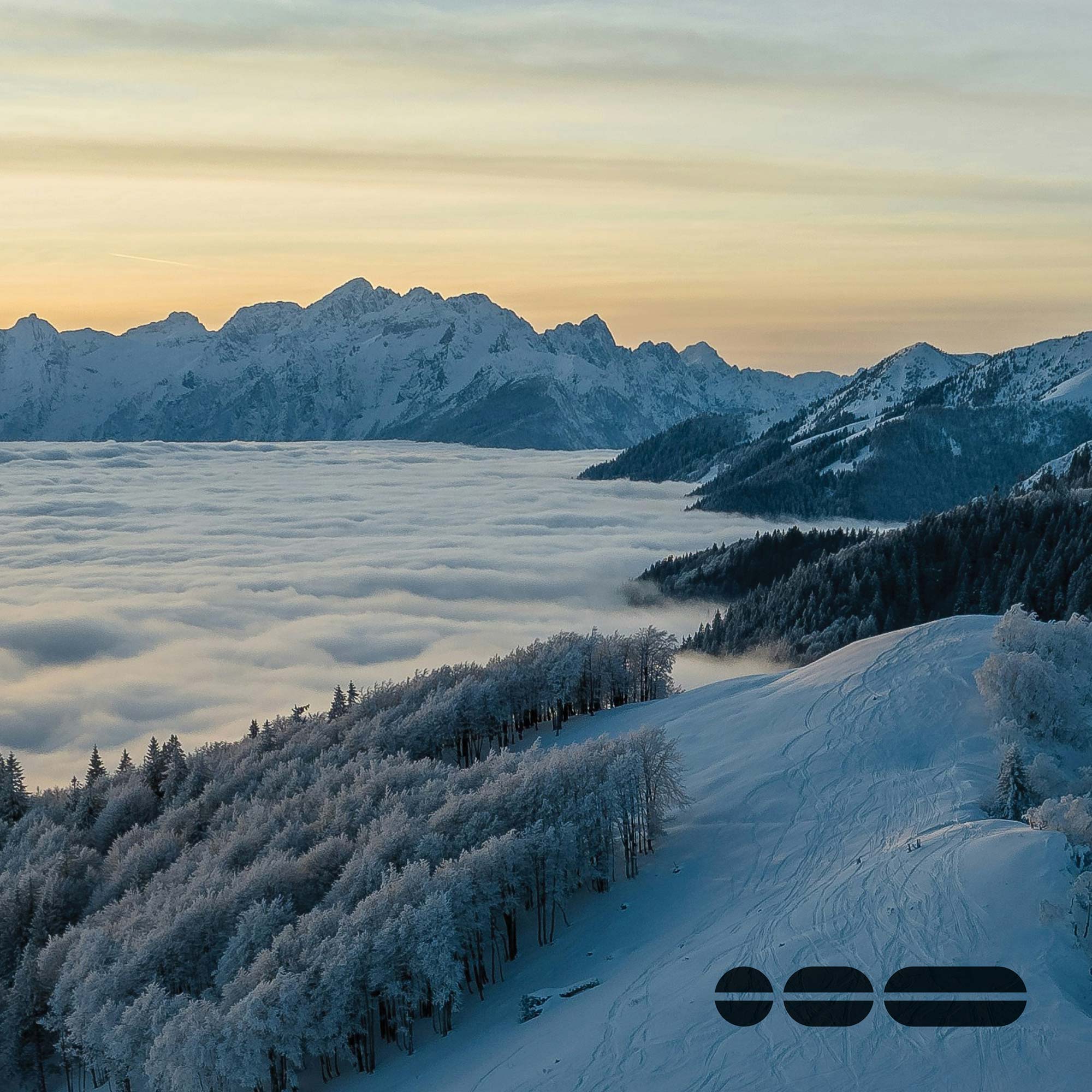 Snow-covered landscape scene with mountains and yellow sunrise in the background, and with snow covered trees and open land in the foreground.