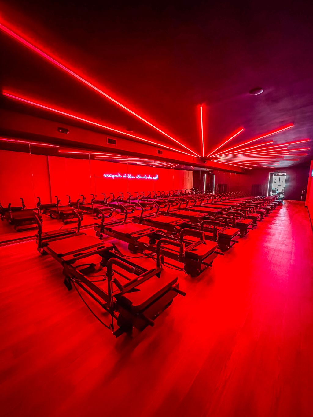 Interior of a fitness studio or gym room with rows of reformer Pilates machines on a wooden floor illuminated by bright red neon ceiling lights and a mirrored wall reflecting the equipment.