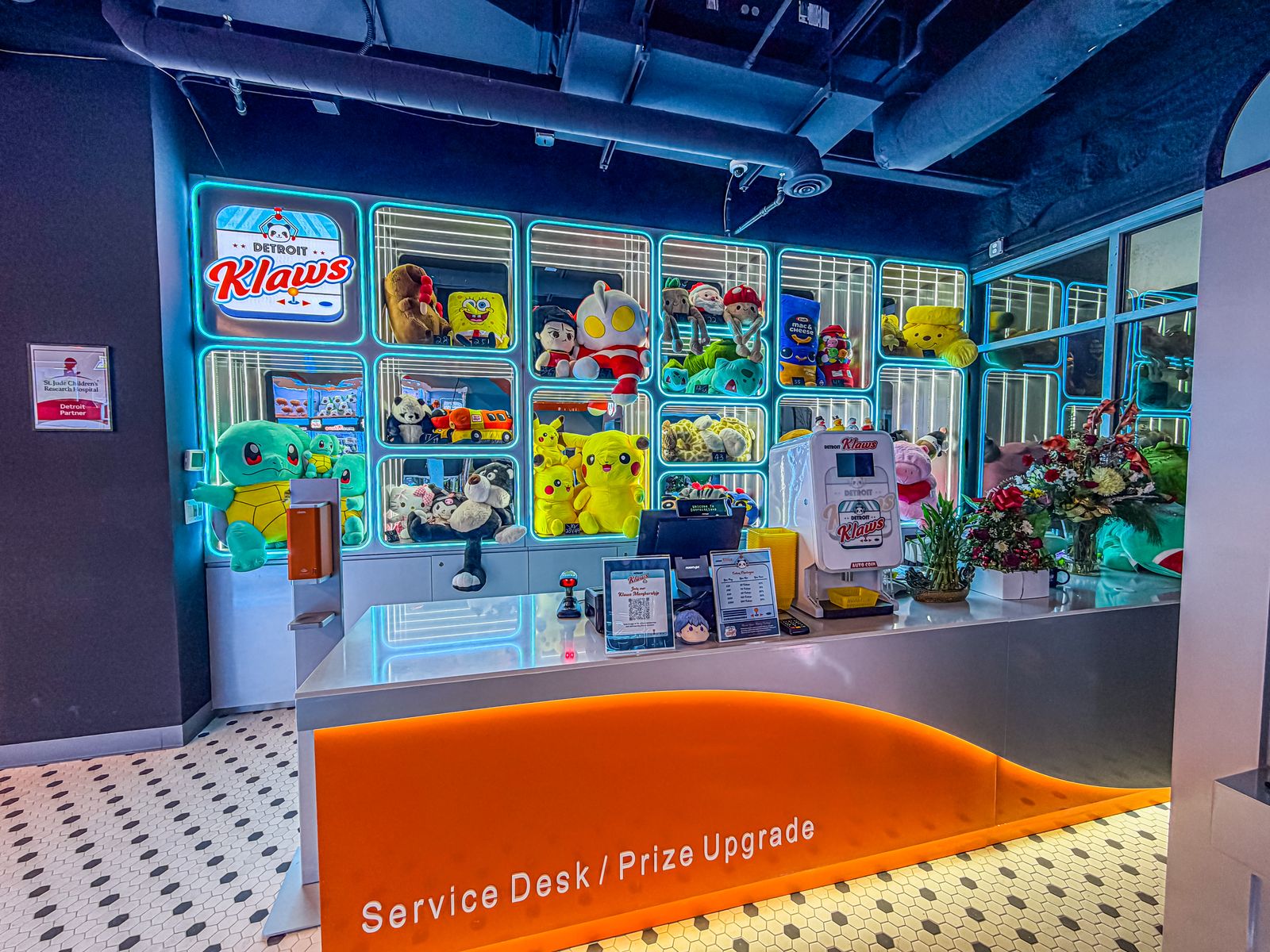 Service desk at Detroit Klaws arcade with an orange front panel labeled 'Service Desk / Prize Upgrade,' a touchscreen register, and a white dispenser machine, backed by shelves illuminated with blue neon lights displaying various stuffed toys including Pikachu, SpongeBob, Ultraman, Squirtle, and other plush characters, alongside flowers and plants on the counter.
