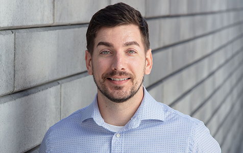 Smiling young man with short dark hair and light beard wearing a light blue button-up shirt standing against a grey tiled wall.