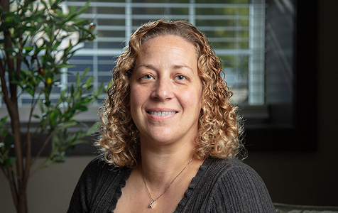 Woman with curly blond hair wearing a dark sweater and a necklace, smiling in an indoor setting with a plant and window blinds in the background.